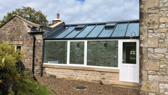 A modern conservatory in an old farm style home with black frames on the glass roof and a white door.