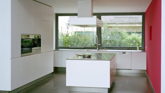Modern white kitchen with a white island in the centre of the room and double aperture aluminium windows looking out onto a hedge.